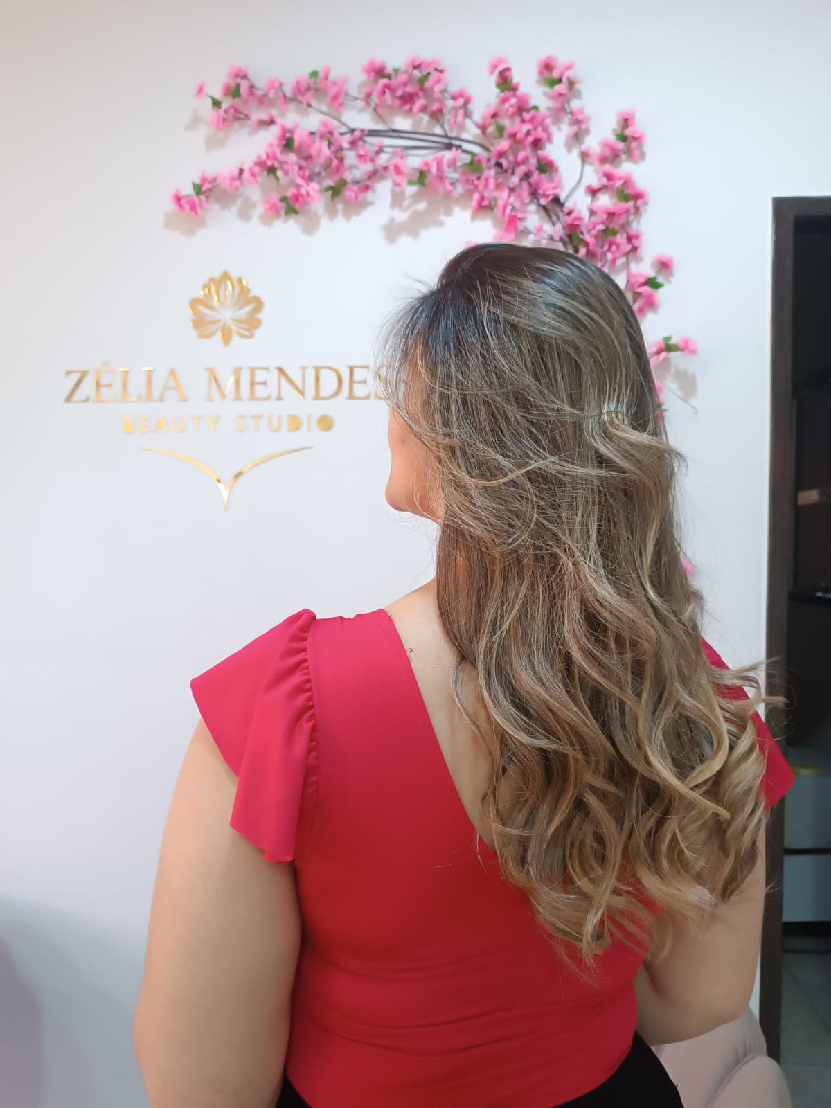 Mulher de costas com cabelos longos, ondulados e com luzes, vestindo uma blusa vermelha.
