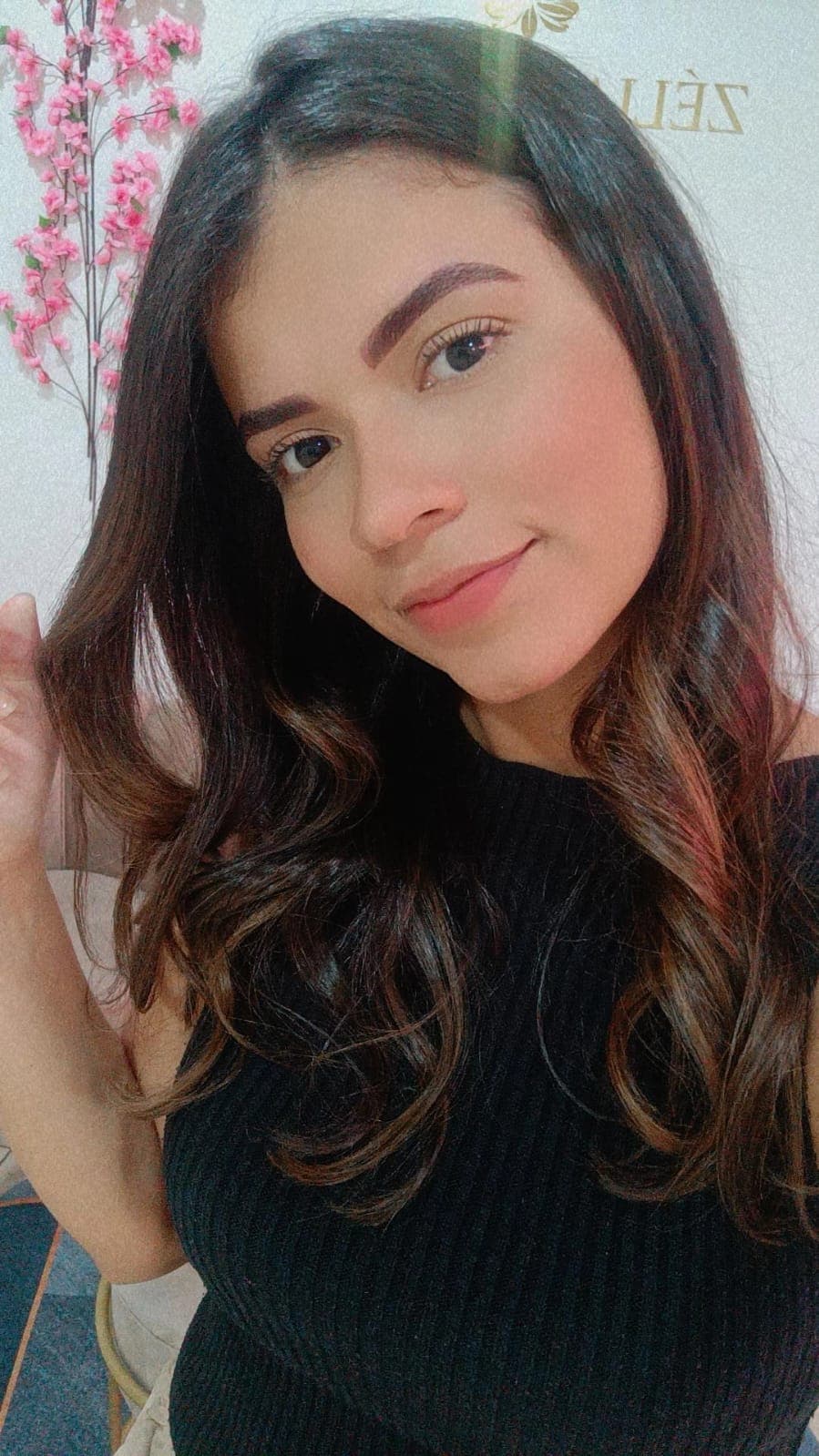 Mulher jovem com longo cabelo escuro ondulado e blusa preta sorrindo para a câmera.