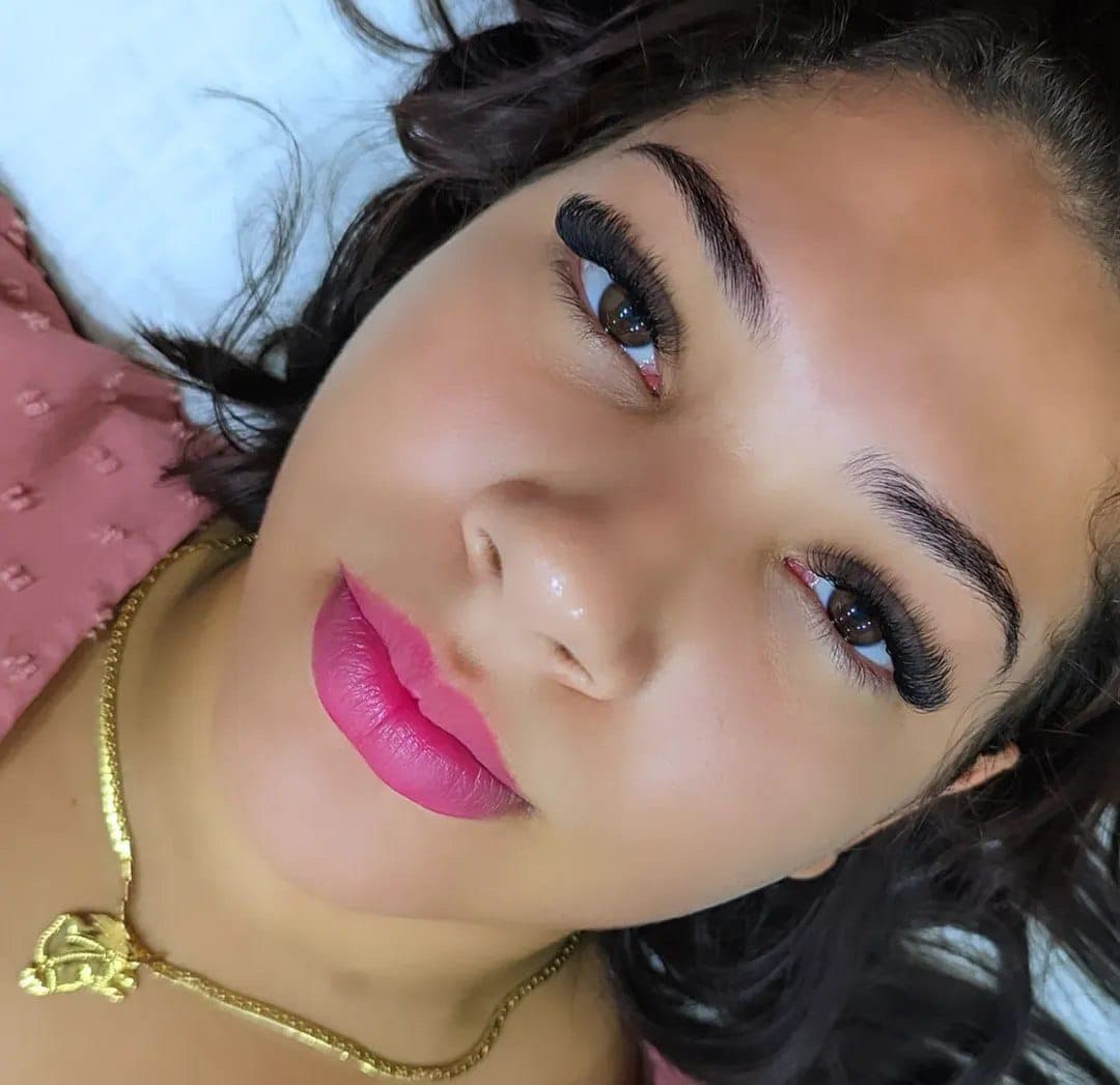 Mulher deitada com extensões de cílios volumosas, sobrancelhas definidas, batom rosa e colar dourado.
