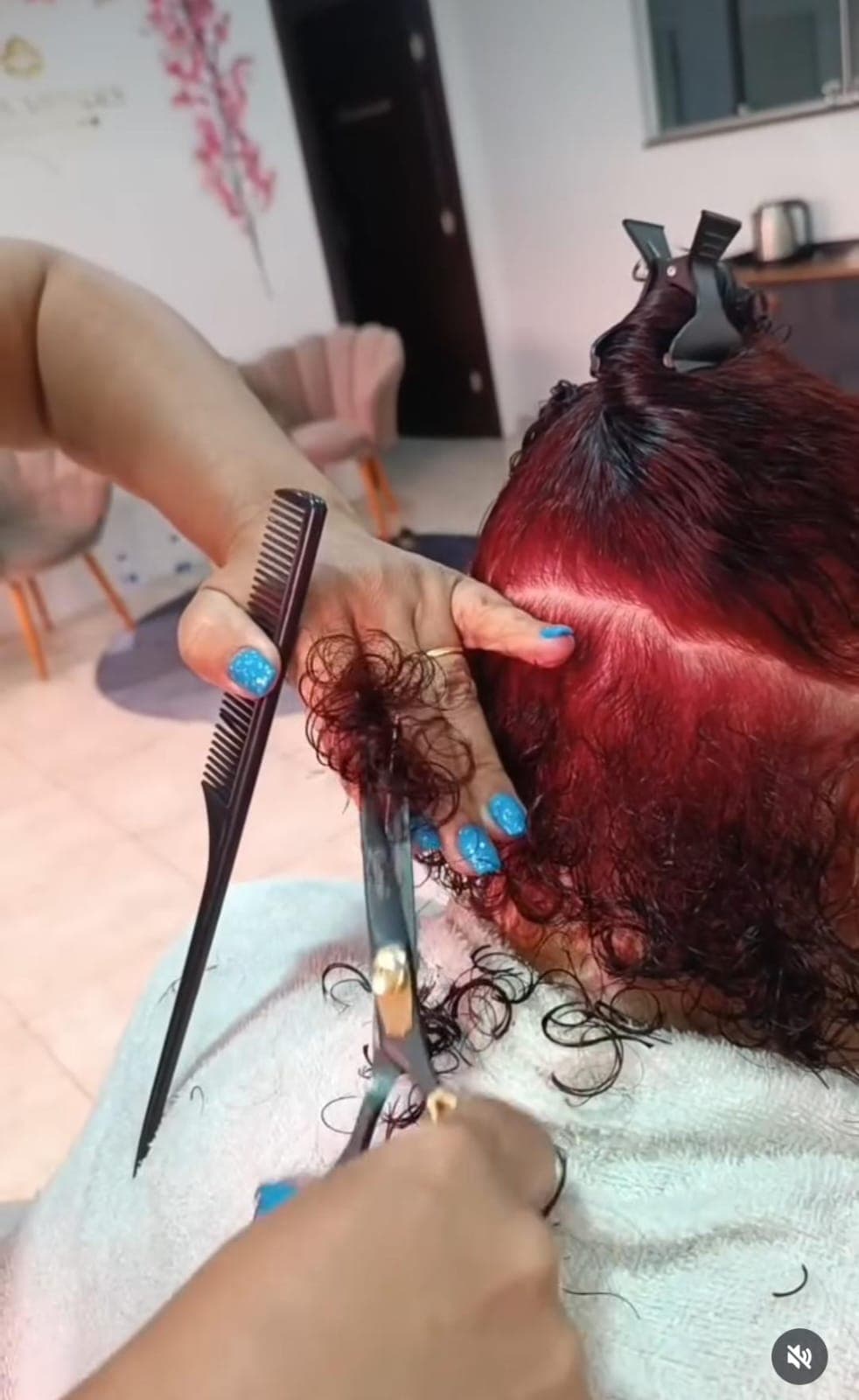 Cabeleireira com unhas azuis corta mecha de cabelo cacheado vermelho usando tesoura e pente preto.